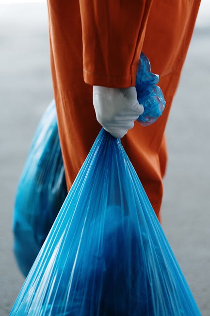 A worker in a blue uniform and an orange safety vest is placing a blue wheeled rubbish bin into the back of a white waste collection truck parked alongside a narrow cobblestone street. The truck's rea