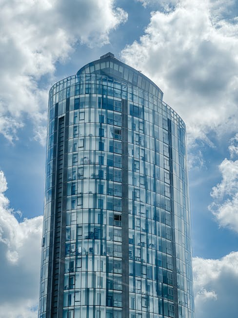 A tall, modern glass commercial building with a curved, cylindrical shape, reflecting the cloudy sky above. The building's exterior is composed of numerous rectangular glass panels arranged in a grid
