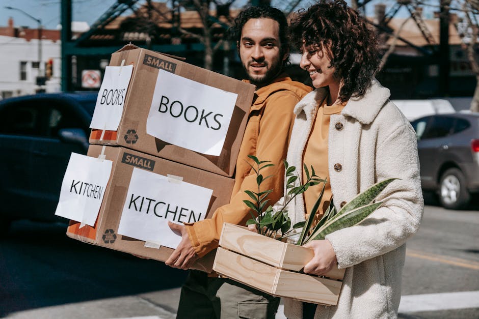 A man and a woman are outside on a street during daytime, preparing for a home relocation. The man is holding two cardboard boxes labeled 'BOOKS' and 'KITCHEN,' both securely taped and stacked, with white labels indicating their contents. The woman is carrying a wooden box with green houseplants, including leafy foliage, wrapped with clear plastic to protect them during transport. Behind them, parked cars and a residential street are visible, under a partly cloudy sky. The scene captures the loading process typical of professional removals by Man With a Van Brentford, emphasizing careful packing and transport of household items, including books, kitchenware, and plants, in preparation for a move to a new property. The image highlights aspects of packing and logistics involved in street-by-street house moving services, visually supporting the concepts of furniture transport and moving organization.