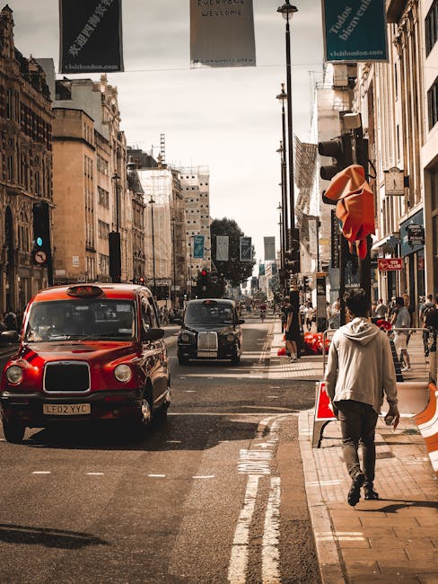 The image shows a busy city street scene with a red traditional London taxi cab and another black vehicle waiting at a pedestrian crossing. The street is flanked by tall, historic buildings with various shopfronts and signage. Overhead, several hanging banners promote local events and businesses, and streetlights are mounted on tall poles. Pedestrians are visible on the sidewalk, including a man dressed in a light-colored hoodie walking away from the camera near a small street closure barrier with orange and white cones. Part of a shop-front with red fabric or signage can be seen on the right side. The street appears to be in a central urban area, likely popular for home relocations and furniture transport, with ample space for loading and unloading goods associated with house removals. Overall, the scene captures a typical city environment suitable for logistics related to moving services from [COMPANY_NAME] in the context of residential relocations in Brentford, TW8.