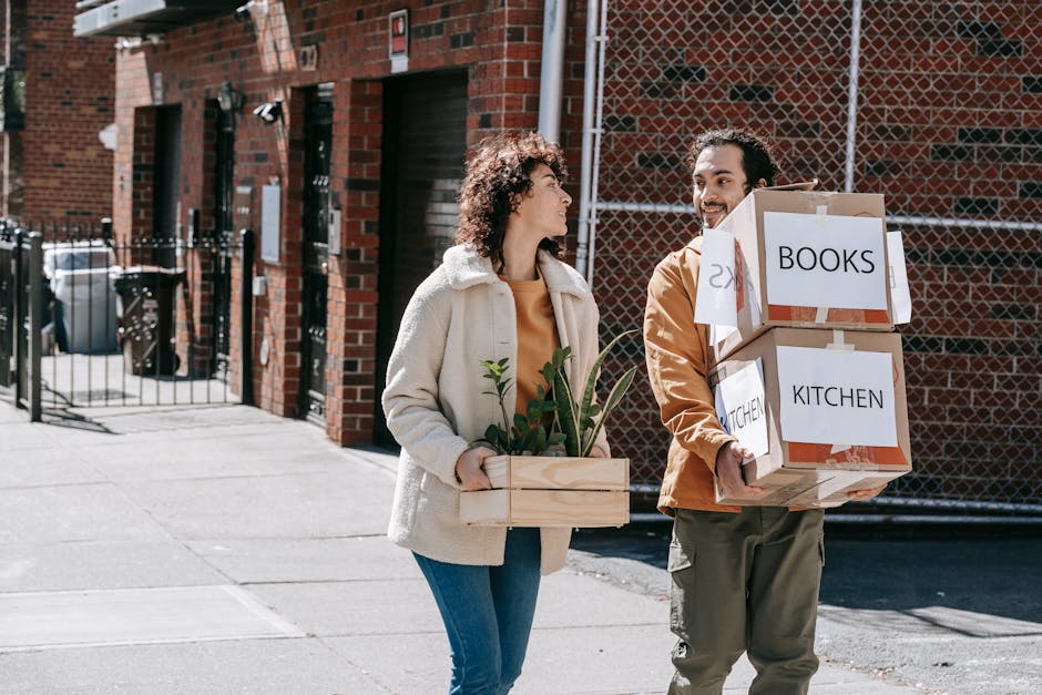 A man and a woman are outside on a street during daytime, preparing for a home relocation. The man is holding two cardboard boxes labeled 'BOOKS' and 'KITCHEN,' both securely taped and stacked, with white labels indicating their contents. The woman is carrying a wooden box with green houseplants, including leafy foliage, wrapped with clear plastic to protect them during transport. Behind them, parked cars and a residential street are visible, under a partly cloudy sky. The scene captures the loading process typical of professional removals by Man With a Van Brentford, emphasizing careful packing and transport of household items, including books, kitchenware, and plants, in preparation for a move to a new property. The image highlights aspects of packing and logistics involved in street-by-street house moving services, visually supporting the concepts of furniture transport and moving organization.