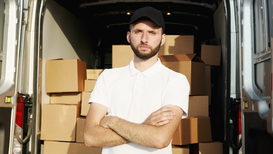 A young man with a beard and serious expression, wearing a black cap and a white polo shirt, standing with arms crossed inside an open moving van. Behind him are stacked cardboard boxes of varying siz
