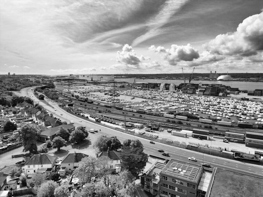 A black and white aerial photograph showing a large urban industrial area with numerous warehouse buildings and shipping containers near a body of water. In the foreground, residential houses with pitched roofs and surrounding trees are visible, along with a curved road lined with parked and moving cars. In the distance, several cranes and construction equipment can be seen, indicating ongoing development, while the sky features scattered clouds. This scene captures a busy port and nearby neighbourhood, appropriate for illustrating logistics, home relocation, or moving services such as those provided by Man With a Van Brentford, with a focus on the transportation and loading process involved in house removals or furniture transport.