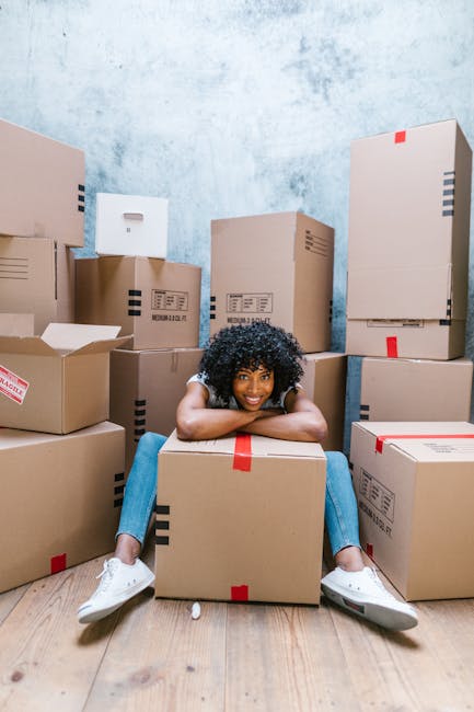 A young woman with curly black hair, wearing a light-colored T-shirt, jeans, and white sneakers, is sitting on the wooden floor inside a room filled with numerous cardboard boxes of various sizes, some sealed with red or black tape and others partially open, revealing packing materials or contents. She is smiling at the camera with her arms resting on a large box in front of her, with her legs spread out and feet flat on the floor. The boxes are stacked against the light blue, textured wall and on the floor, indicating an ongoing home relocation process managed by Man With a Van Brentford. The scene illustrates the packing and loading stages of a furniture transport or house removal, with the boxes prepared for transportation either within a property or onto a vehicle outside, supported by the presence of the boxes and the indoor setting.